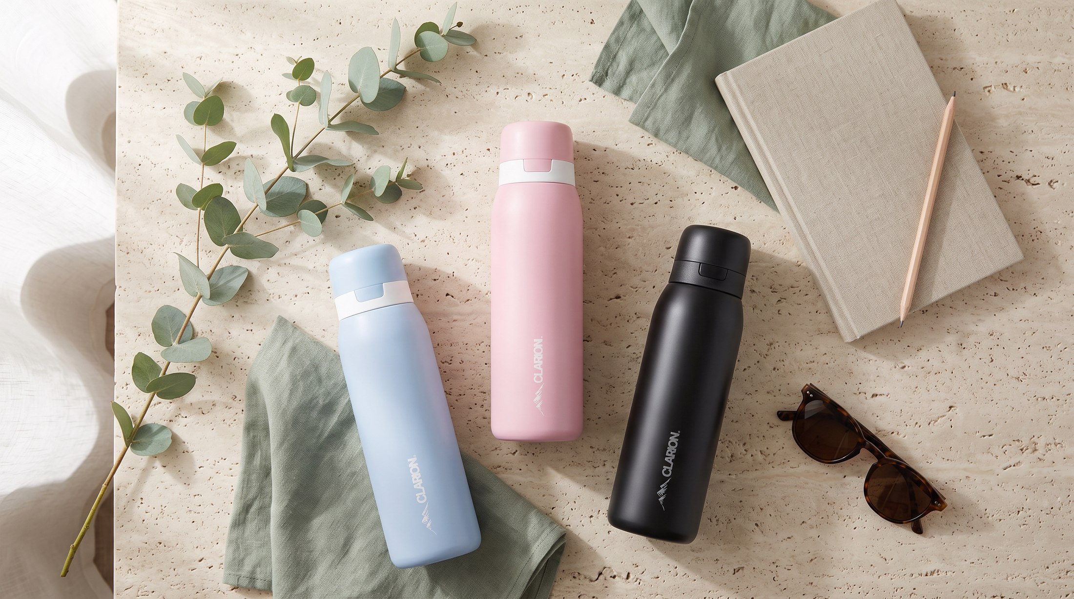 Blue, pink, and black Clarion Bottles laid out on a stone surface with eucalyptus and linen